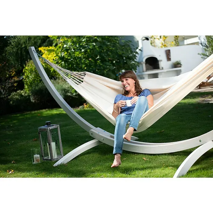 La Siesta Hängematte Brisa Kingsize - L x B: 400 x 180 cm, Vanilla - Eine Frau entspannt in einer Hängematte mit Gestell im Garten und genießt einen Drink. Die Hängematte bietet eine komfortable Ruhezone im Freien.