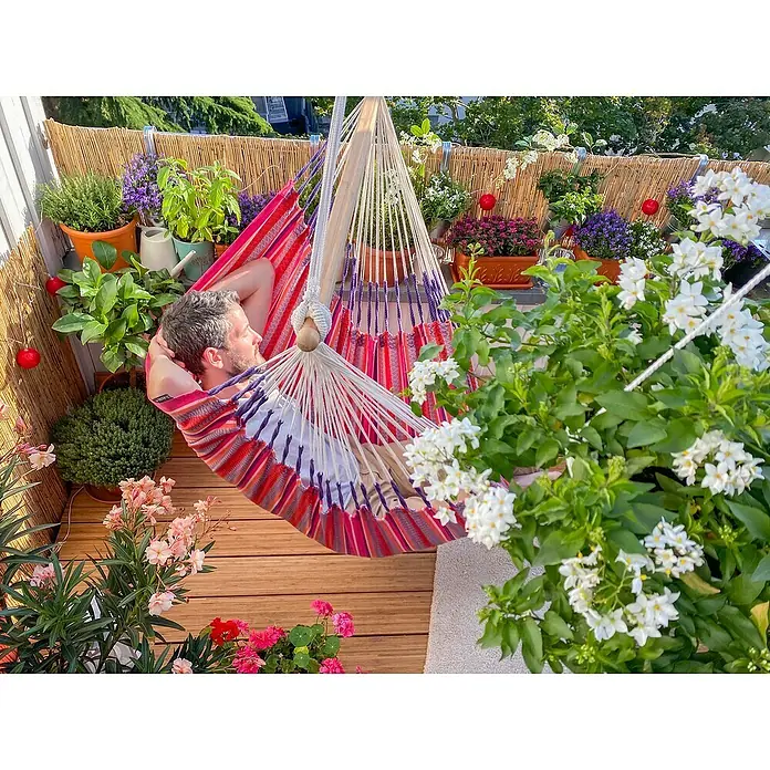 La Siesta Hängesessel Habana Comfort - L x B x H: 180 x 115 x 160 cm, Flamingo - Ein Mann entspannt in einer bunten Hängematte auf einem sonnigen Balkon, umgeben von üppigen Pflanzen und Blumen.