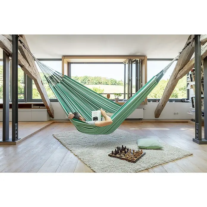 La Siesta Hängematte Modesta - L x B: 400 x 180 cm, Agave, Kingsize - Ein Mann entspannt in einer grünen Hängematte mit Buch über einem Schachbrett auf einem hellen Teppich in einem stilvollen Raum.