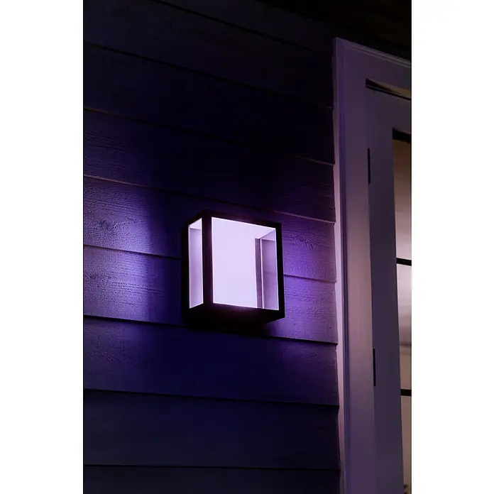 Philips Hue LED-Außenwandleuchte Impress - 8 W, 11,7 x 19 x 24 cm, Schwarz, IP44 - Eine quadratische Außenleuchte in Schwarz strahlt violettes Licht an einer Wand neben einer Tür.