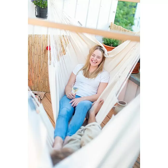 La Siesta Hängesessel Habana Kingsize - L x B x H: 210 x 130 x 170 cm, Latte - Eine lachende Frau entspannt in einer weißen Stoffhängematte auf einem Balkon, ein Weinglas haltend. Füße einer weiteren Person sind sichtbar.