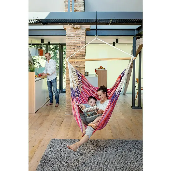 La Siesta Hängesessel Habana Kingsize - L x B x H: 210 x 130 x 170 cm, Flamingo - Eine Mutter und ihr Kind entspannen sich in einer Hängematte im Haus, während der Vater in der Küche kocht. Die Hängematte ist farbenfroh und lädt zum Verweilen ein.