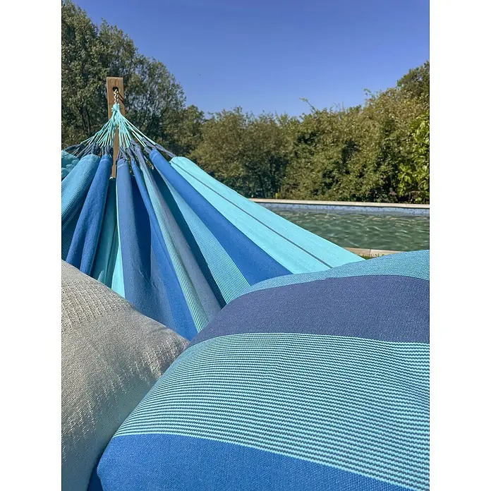 La Siesta Hängematte Modesta - L x B: 350 x 160 cm, Lagoon, Double - Nahaufnahme einer blau gestreiften Hängematte mit Kissen, die zum Entspannen im Freien einlädt, mit Pool und Bäumen im Hintergrund.