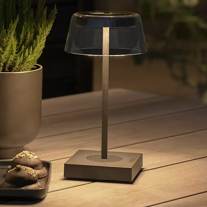 Konstsmide Akku-Tischleuchte Scilla - 2,5 W, L x B x H: 11 x 11 x 27 cm, Schwarz, Warmweiß - Dunkelgraue tragbare Tischleuchte mit klarem Schirm, spendet warmes Licht auf einem Holztisch im Freien.