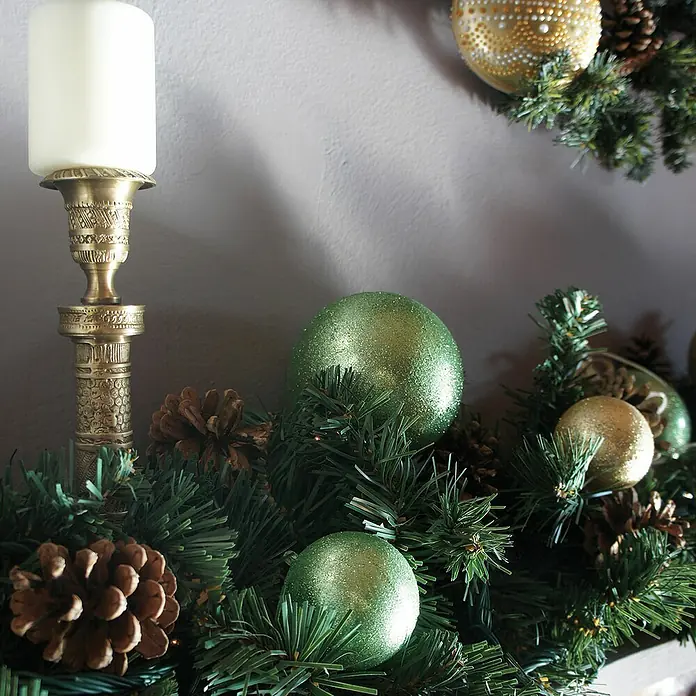 Fleur Purpurina Glitter - Jade Green, 90 g, Brillante - Decoración navideña con una vela en un candelabro dorado, bolas verdes y doradas y piñas en una guirnalda verde.