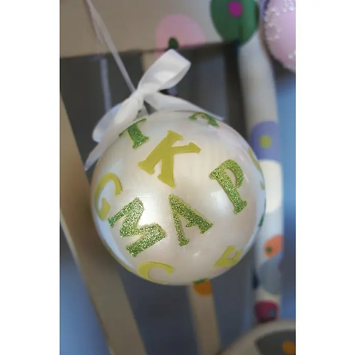 Fleur Purpurina Glitter - Jade Green, 90 g, Brillante - Primer plano de un adorno navideño blanco con letras verdes y amarillas, colgado de una cinta blanca, evocando un ambiente festivo.