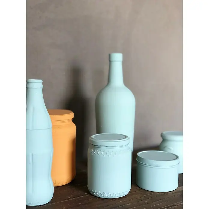 Fleur Pintura decorativa en spray Chalky Look - Malmo, Mate, 300 ml - Recipientes de vidrio pintados en varias formas y colores dispuestos sobre una superficie de madera con un fondo simple.