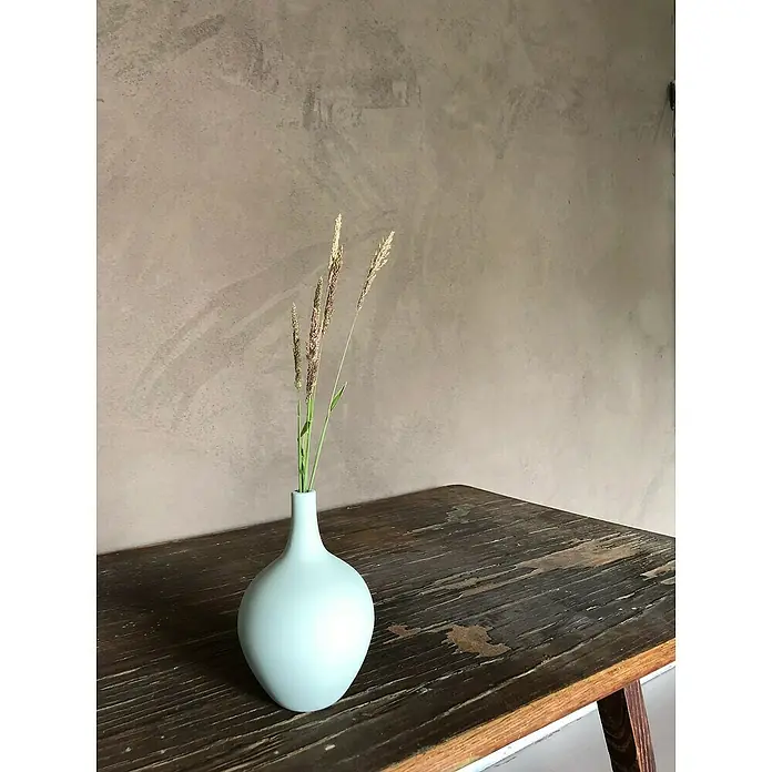 Fleur Pintura decorativa en spray Chalky Look - Malmo, Mate, 300 ml - Elegante jarrón azul claro con hierba delicada sobre una mesa rústica de madera, con una pared lisa que enfatiza la estética minimalista.