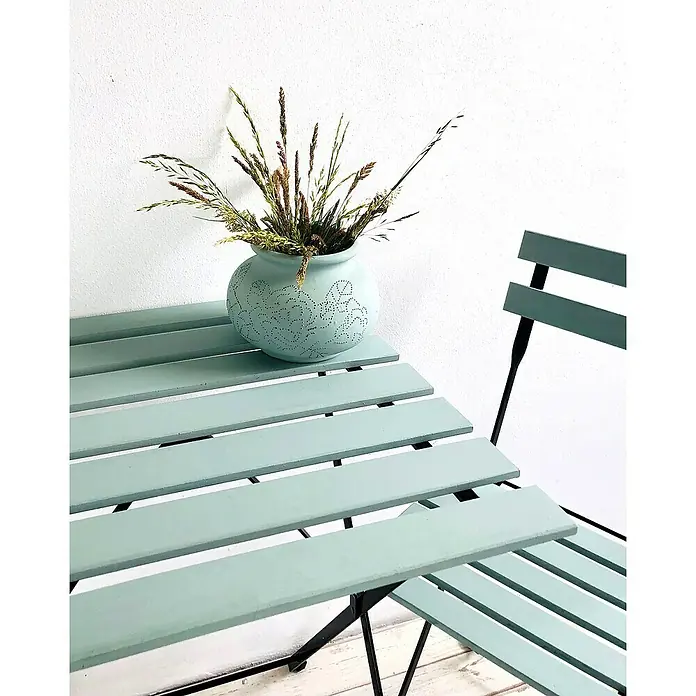Fleur Pintura decorativa en spray Chalky Look - Malmo, Mate, 300 ml - Primer plano de una mesa de jardín turquesa con una silla y un jarrón adornado con hierbas, que irradia una tranquilidad acogedora.