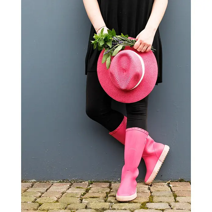 Fleur Pintura decorativa en spray Chalky Look - Penelope, Mate, 300 ml - Primer plano de una persona con botas de agua que sostiene un sombrero rosa y hierbas frente a una pared gris.