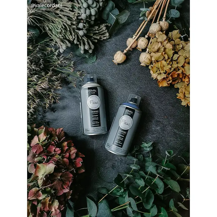 Fleur Pintura decorativa en spray Chalky Look - Smoky, Mate, 300 ml - Dos botes de spray de pintura Fleur Chalky Look Designer entre arreglos florales sobre un fondo oscuro.