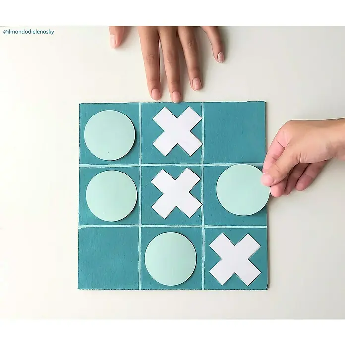 Fleur Pintura decorativa en spray Chalky Look - Capetown Azul, Mate, 300 ml - Vista aérea de un juego de Tic-Tac-Toe con tablero azul, círculos verde claro y símbolos X blancos, donde una persona está colocando un círculo.