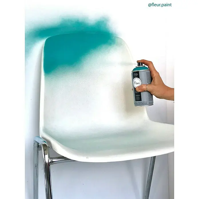 Fleur Pintura decorativa en spray Chalky Look - Istanbul, Mate, 300 ml - Una silla blanca está siendo rociada con pintura en aerosol turquesa Fleur, creando un elegante degradado de color. Ideal para proyectos DIY.