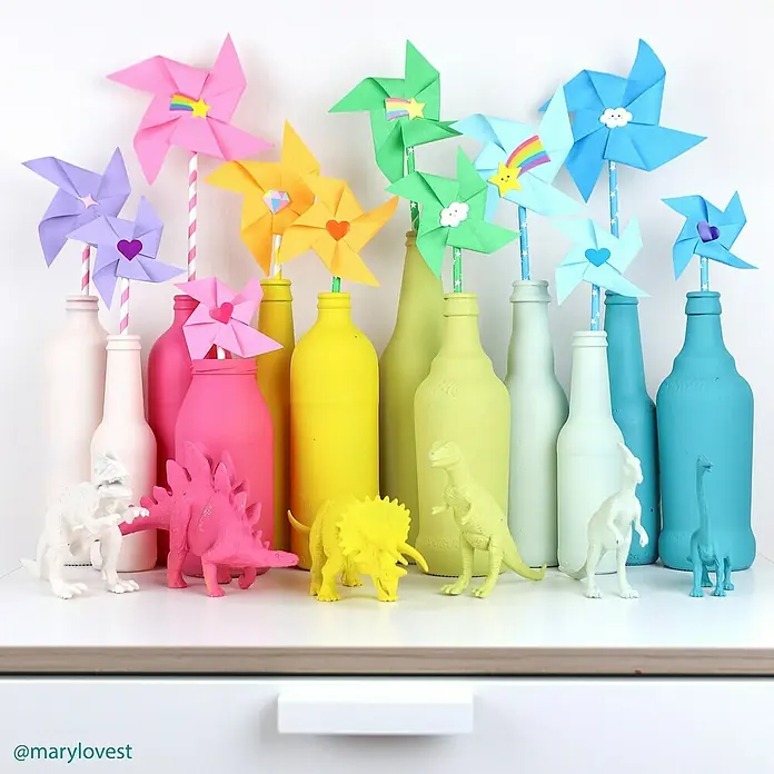 Fleur Pintura decorativa en spray Chalky Look - Istanbul, Mate, 300 ml - Coloridos molinetes de papel en botellas pintadas con dinosaurios de juguete a juego dispuestos en un estante blanco.