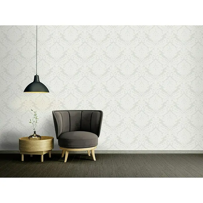 A.S. Création Vliestapete Flock - Metallic/Weiß, Ornament, 10,05 x 0,53 m - Elegantes Interieur mit einem grauen Sessel und einem Beistelltisch unter einer modernen Pendelleuchte vor einer verzierten Tapete.