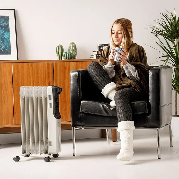 Taurus Ölradiator New Dakar - 2.000 W, Raumgröße bis ca. 20 m², Weiß - Eine Frau sitzt in einem Sessel neben einem weißen Heizkörper und trinkt eine Tasse. Der Heizkörper sorgt für Wärme und Komfort.