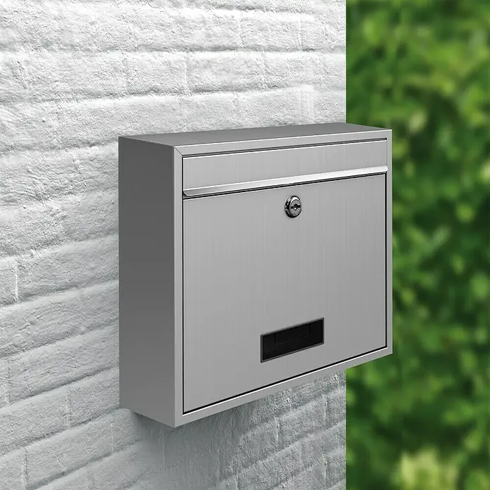Basi Wandbriefkasten BK 900 - L x B x H: 11,6 x 36 x 31,5 cm, Edelstahl, Edelstahl, Namensschild - Ein moderner Briefkasten aus Edelstahl an einer weißen Ziegelwand. Der Briefkasten verfügt über ein Schloss und einen Schlitz.