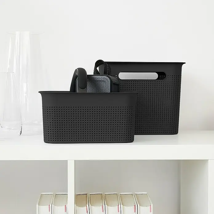 Rotho Aufbewahrungsbox Brisen - L x B x H: 26,2 x 18 x 13 cm, Kunststoff, Anthrazit - Auf dem Regal stehen zwei schwarze Aufbewahrungskörbe mit Löchern. Ein kleiner Lautsprecher steht in dem einen Korb.