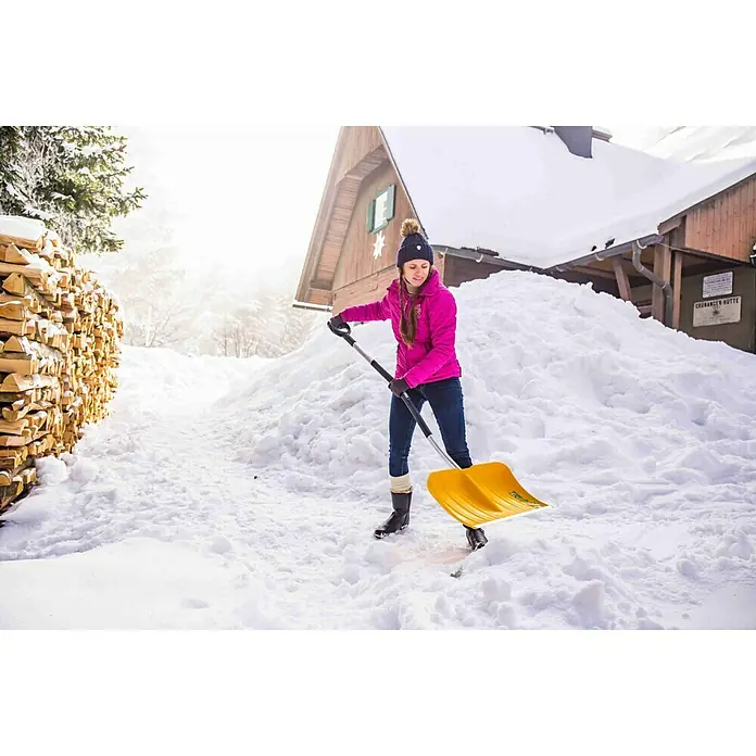 Freund Victoria Schneeschieber - Breite: 53 cm, Ergostiel, Material Stiel: Aluminium - Frau schaufelt Schnee mit einer gelben Schneeschaufel vor einem Holzhaus. Die Schaufel ist gut sichtbar und steht im Fokus.