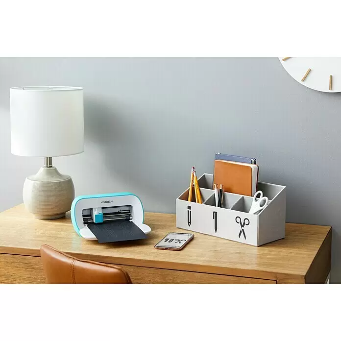 Cricut Joy Schneideplotter Smarte Schneide- und Zeichenmaschine - Türkis/Weiß, Bluetooth, Schnittbreite: 10,4 cm, Schnittlänge: 1,2 m - Eine hellblaue Cricut Joy Maschine auf Holzschreibtisch schneidet schwarze Vinylformen. Ein Telefon zeigt Designs neben einem personalisierten Organizer.