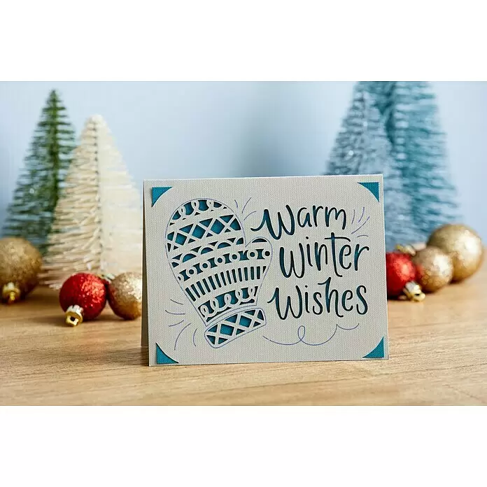 Cricut Joy Schneideplotter Smarte Schneide- und Zeichenmaschine - Türkis/Weiß, Bluetooth, Schnittbreite: 10,4 cm, Schnittlänge: 1,2 m - Eine hellfarbene Wintergrußkarte mit ausgeschnittenem Fäustling und "Warm Winter Wishes"-Text, umgeben von verschwommenen Weihnachtsdekorationen.
