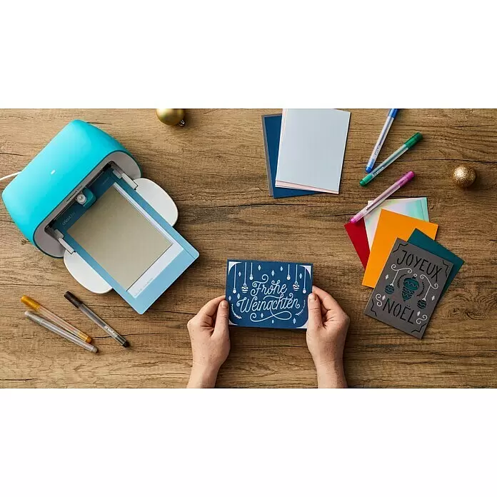 Cricut Joy Schneideplotter Smarte Schneide- und Zeichenmaschine - Türkis/Weiß, Bluetooth, Schnittbreite: 10,4 cm, Schnittlänge: 1,2 m - Eine hellblaue Cricut Joy Maschine auf einem Holztisch, daneben bunte Karten und Stifte. Hände halten eine fertige 'Frohe Weihnachten' Karte.
