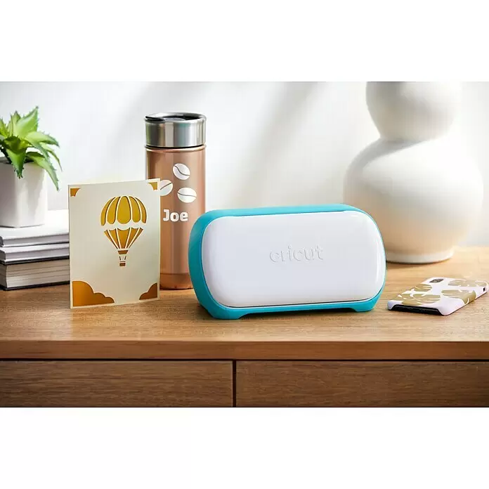 Cricut Joy Schneideplotter Smarte Schneide- und Zeichenmaschine - Türkis/Weiß, Bluetooth, Schnittbreite: 10,4 cm, Schnittlänge: 1,2 m - Eine weiß-blaue Cricut-Maschine steht auf Holz, umgeben von maßgefertigten Artikeln wie Karte, Becher und Handyhülle, die ihre Bastelfähigkeiten zeigen.