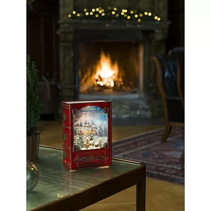 Konstsmide LED-Weihnachtsdekofigur Buch - 13,5 x 19,3 cm - Rote, buchförmige Weihnachtsschneekugel auf einem Tisch in einem gemütlichen Raum mit Kamin.