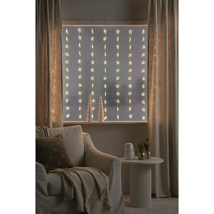 Konstsmide LED-Lichtervorhang Sterne - B x H: 140 x 120 cm, Innen, Kunststoff, Warmweiß - Gemütliche Raumecke mit einem bequemen Sessel, einem Beistelltisch und einer Fensterdekoration mit Sternenlichtern.