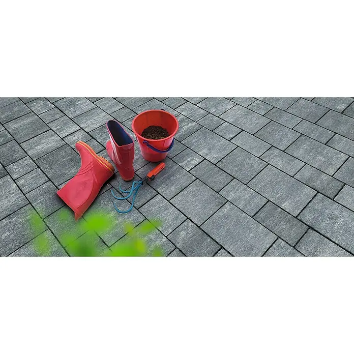 Diephaus Pflasterstein Ventura - Graphit, 19 Stk., Rechteckig - Nahaufnahme von roten Gummistiefeln, einem Eimer mit Erde und einem Rechen auf grauen Pflastersteinen, die einen Gartenbereich verschönern.