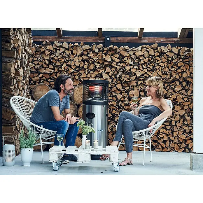 Enders Estufa de exterior Polo - 3 kW - 6 kW, Altura: 115 cm - Un calentador de patio de acero inoxidable calienta a una pareja que se relaja y disfruta de bebidas en un patio con una pared decorativa de leña.