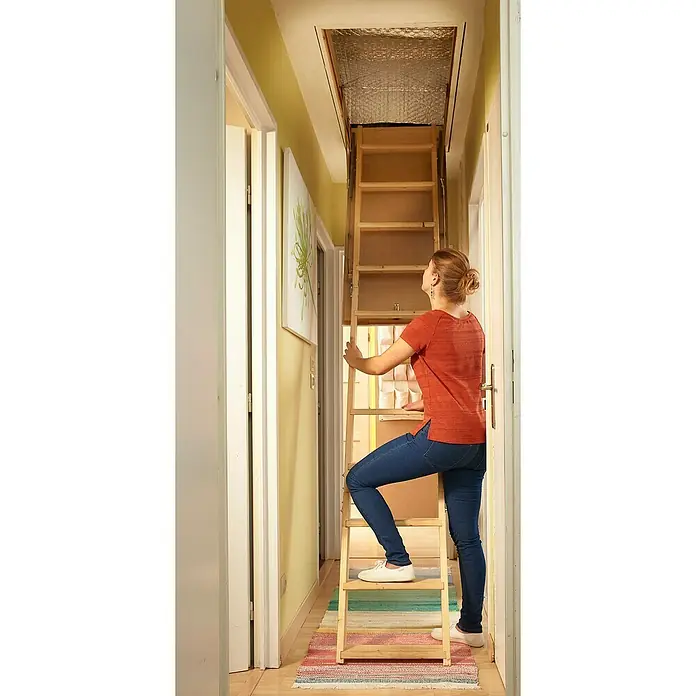 Isolierabdeckung für ausziehbare Bodentreppen - 130 cm x 70 cm x 8 mm - Eine Frau steigt eine Holztreppe hoch zu einer Dachbodenluke, die mit reflektierendem Material isoliert ist. Die Leiter steht auf einem bunten Teppich.