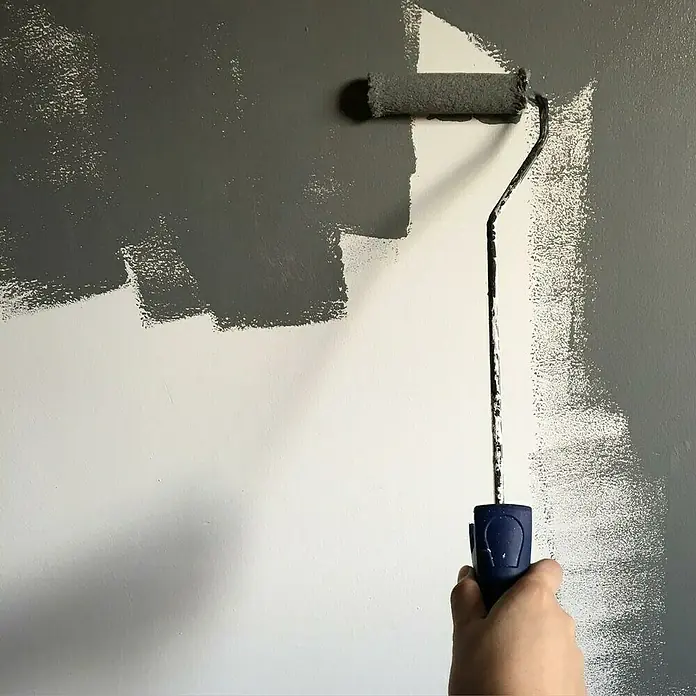 Fleur Esmalte de color Multi-Surface Eggshell - Cream Love, 375 ml, Satinado, Consumo por m²: 12 l - Primer plano de una persona pintando una pared con un rodillo y pintura gris, con foco en la aplicación de la pintura.