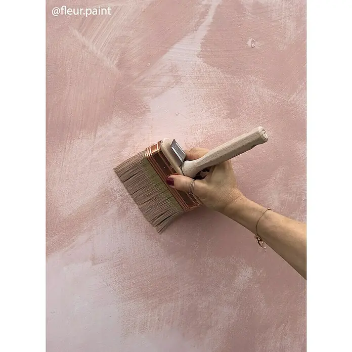 Fleur Pintura para efectos decorativos Lime Paint - Jo, 750 ml, Mate - Primer plano de una persona pintando una pared rosa con una brocha ancha, resaltando la textura y la aplicación de la pintura del pincel.