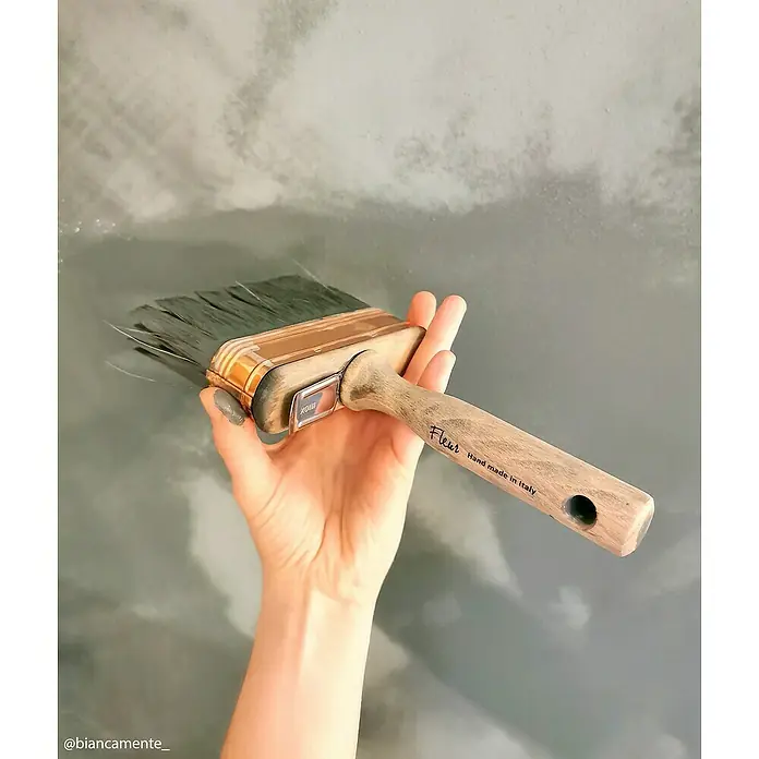Fleur Pintura para efectos decorativos Lime Paint - Woodland Spirit, 750 ml, Mate - Una mano sostiene un pincel Fleur con mango de madera y cerdas grises frente a un fondo pintado de gris.