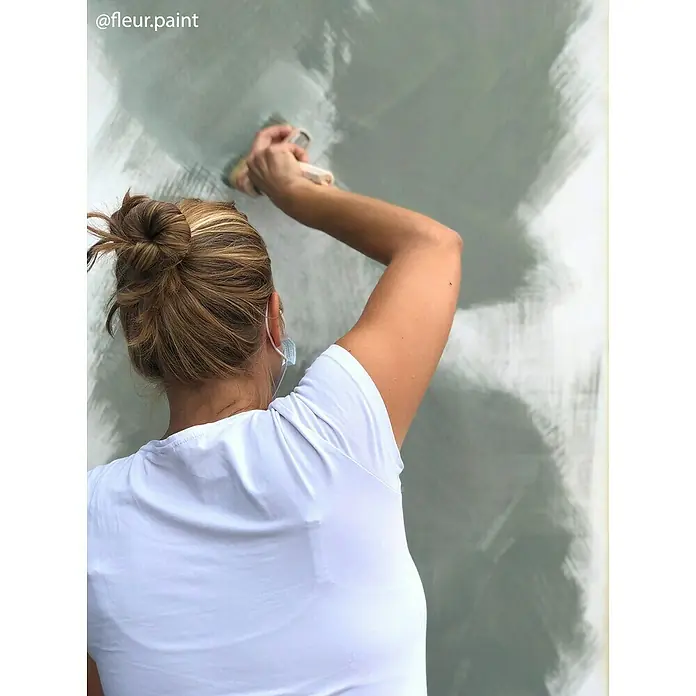Fleur Pintura para efectos decorativos Lime Paint - Soho Green, 2,5 l, Mate - Una mujer con un moño pinta una pared con un pincel, creando una textura única que acentúa la pintura de alta calidad.