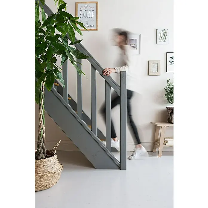 V33 Esmalte de color Renovation Perfection suelos de cerámica - Marrón topo, Satinado, 2 l - Una mujer sube una escalera de madera gris junto a una planta en maceta en una cesta, creando un ambiente acogedor.