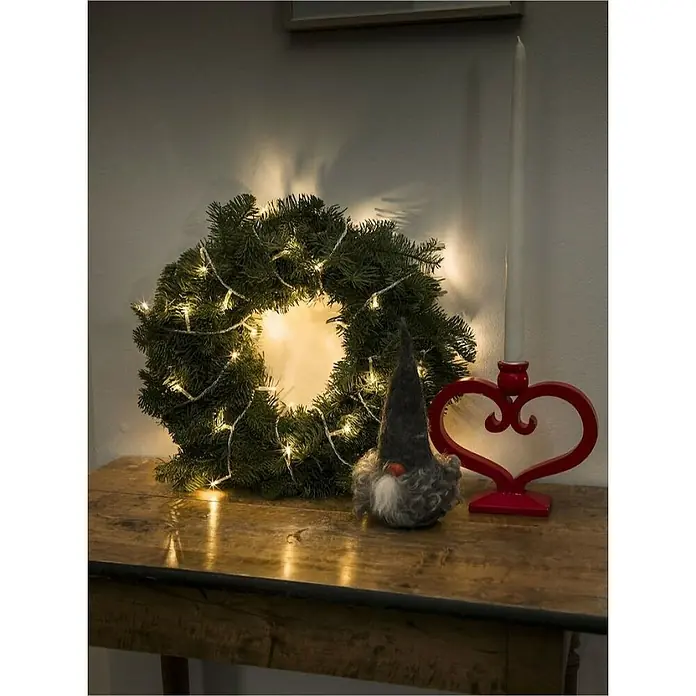 Konstsmide LED-Lichterkette - Innen, Lichtfarbe: Warmweiß, Anzahl Leuchtmittel: 35 Stk., Netzbetrieben, Transparent - Festlicher Weihnachtsschmuck: Ein beleuchteter Kranz, ein Wichtel und ein roter Kerzenständer in Herzform auf einem rustikalen Tisch.