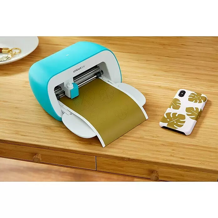 Cricut Joy Schneideplotter Starter Bundle - Türkis/Weiß, Bluetooth, Schnittbreite: 10,4 cm, Schnittlänge: 1,2 m - Eine türkis-weiße Cricut Joy Maschine schneidet goldfarbenes Material mit Monstera-Blattformen auf einem Holztisch, daneben ein Handy mit passender Hülle.
