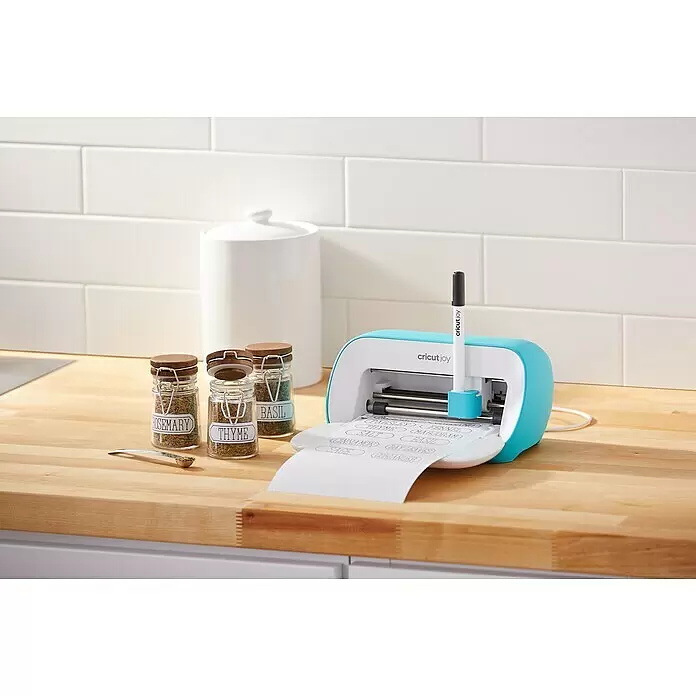 Cricut Joy Schneideplotter Starter Bundle - Türkis/Weiß, Bluetooth, Schnittbreite: 10,4 cm, Schnittlänge: 1,2 m - Eine teal-weiße Cricut Joy Maschine druckt Gewürzglasetiketten auf einer Holztheke neben beschrifteten Gläsern.