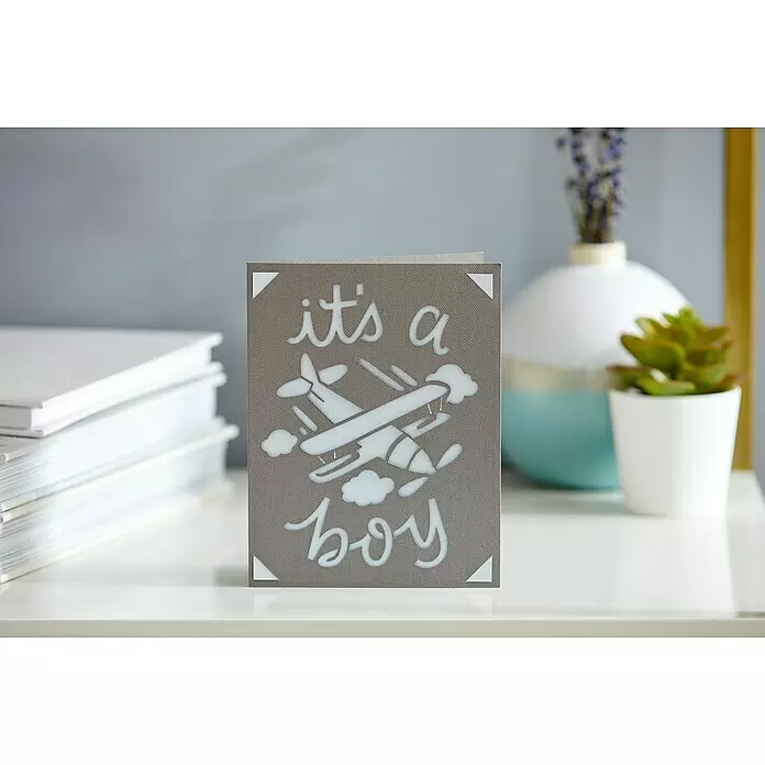 Cricut Joy Schneideplotter Starter Bundle - Türkis/Weiß, Bluetooth, Schnittbreite: 10,4 cm, Schnittlänge: 1,2 m - Eine graue Babypartykarte mit "it's a boy" und einem Flugzeug-Stanzmotiv, auf einer weißen Oberfläche stehend.