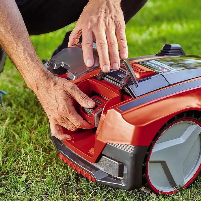 Einhell PXC 18V Mähroboter FREELEXO CAM 500 - 500 m², Schnittbreite: 18 cm, Kamera-gestützte Navigation - Ein Mann, der den Akku eines Einhell-Mähroboters einsetzt, wobei die rote Farbe des Mähers und die grüne Umgebung hervorstechen.