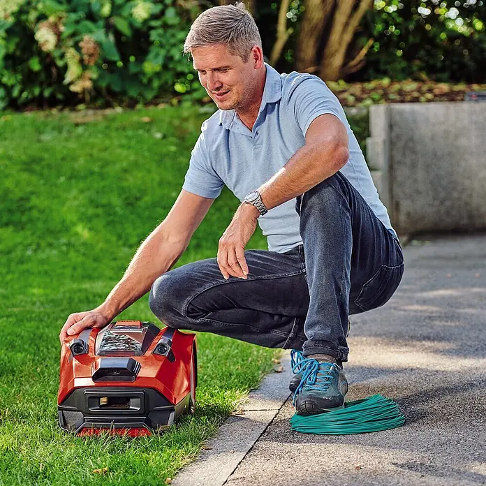 Einhell PXC 18V Mähroboter FREELEXO CAM 500 - 500 m², Schnittbreite: 18 cm, Kamera-gestützte Navigation - Mann hockt neben einem roten Mähroboter und einem grünen Kabel auf einer Rasenfläche.
