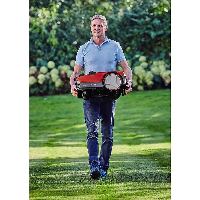 Einhell PXC 18V Mähroboter FREELEXO CAM 500 - 500 m², Schnittbreite: 18 cm, Kamera-gestützte Navigation - Ein Mann trägt einen roten Mähroboter über einen Rasen, umgeben von Bäumen. Der Rasenroboter ist das Produkt im Fokus.