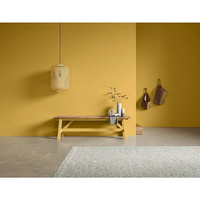 SCHÖNER WOHNEN-Farbe Naturell Wandfarbe Kreidefarbe - Blütenzauber - warmes Gelb, Matt, 2,5 l, Konservierungsmittelfrei - Einrichtungsszene mit gelbem Wandhintergrund, einer Holzbank, einer Hängelampe und einem hellen Teppich im Vordergrund.