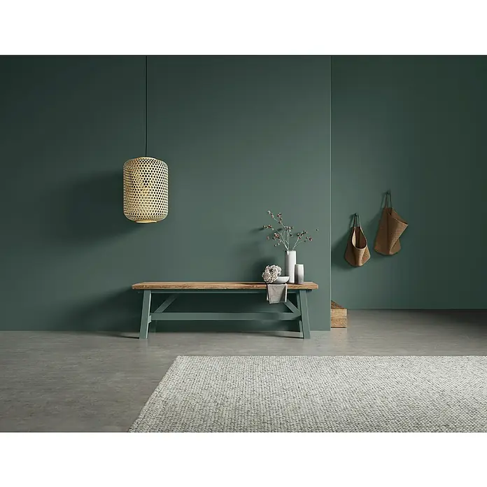 SCHÖNER WOHNEN-Farbe Naturell Wandfarbe Kreidefarbe - Waldgeflüster - tiefes Naturgrün, Matt, 2,5 l, Konservierungsmittelfrei - Einrichtungsszene mit einer Pendelleuchte, einer Bank mit Deko und einem Teppich in einem Raum mit grünen Wänden.