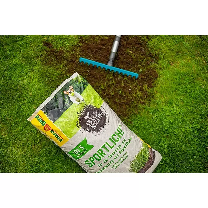 Floragard Rasenerde Sportlich - 40 l, Torffrei - Ein Sack Flora gard Bio-Erde SPORTLICH! Rasenerde liegt auf grünem Gras neben einer frisch geharkten Erdfläche und einem blauen Rechen.