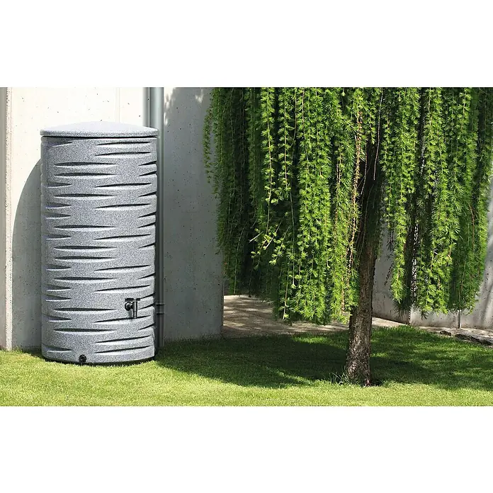 Arves Regenspeicher Freze - 290 l, Granit - Grauer Regenwassertank mit dekorativer Oberfläche und einem Wasserhahn vor einer Betonwand, neben einem grünen Baum auf Rasen.