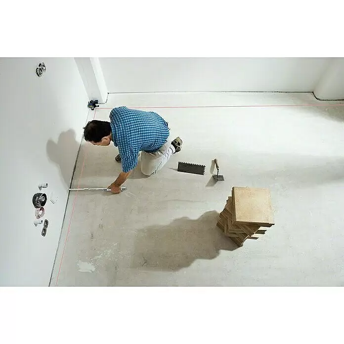 Bosch Professional Fliesenlaser GTL 3 - Max. Arbeitsbereich: 20 m, 4 -tlg. - Mann bereitet einen Boden mit Laserwasserwaage und Messwerkzeug für Fliesenarbeiten vor. Gestapelte Fliesen, Zahnspachtel und Kelle liegen bereit.