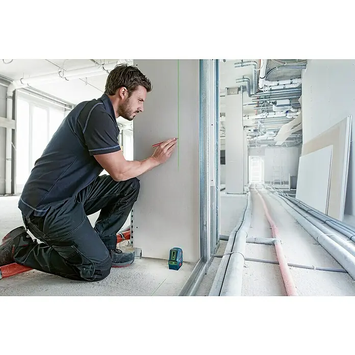 Bosch Professional Linienlaser GLL 2-10 - Max. Arbeitsbereich: 10 m, 3 -tlg. - Ein Bauarbeiter kniet neben einem Bosch-Linienlaser GLL 2-15 G, der eine grüne Linie an eine Wand projiziert, während er mit einem Stift Markierungen setzt.